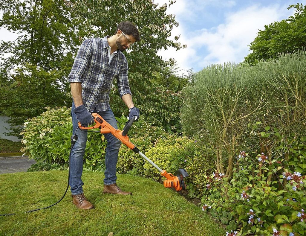 Dans son jardin se trouve un homme avec une chemise à carreaux et un pantalon, utilisant un rotofil électrique.