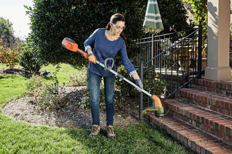 Une femme habillée en bleu porte une débroussailleuse sur batterie. Elle est devant son jardin et à côté des escaliers qui mènent à sa maison.