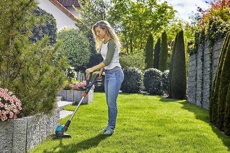 Au milieu de son jardin se trouve une jeune femme qui débroussaille proche d'un gros pot d'arbuste avec le reste du jardin en fond.