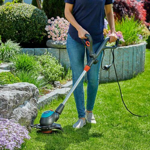 Une femme vêtue de bleu utilise une débroussailleuse électrique avec fil. Elle se trouve dans un jardin qui comprend un décor varié derrière elle.