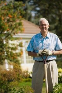 Un home âgé en chemise bleue et pantalon beige pose devant sa maison et son jardin qui se trouve au deuxième plan.