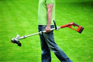 Un homme tient sa débroussailleuse à batterie dans la main. Celui-ci a un t-shirt vert et un jean. Derrière lui se trouve un jardin plat.