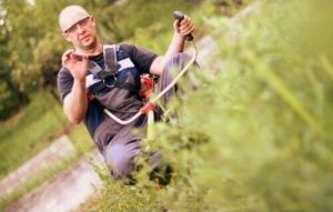 En premier plan, on voit de l'herbe haute floutée. EN second plan, un homme, équipé de sa débroussailleuse, nous regarde et nous fait un signe de la main.